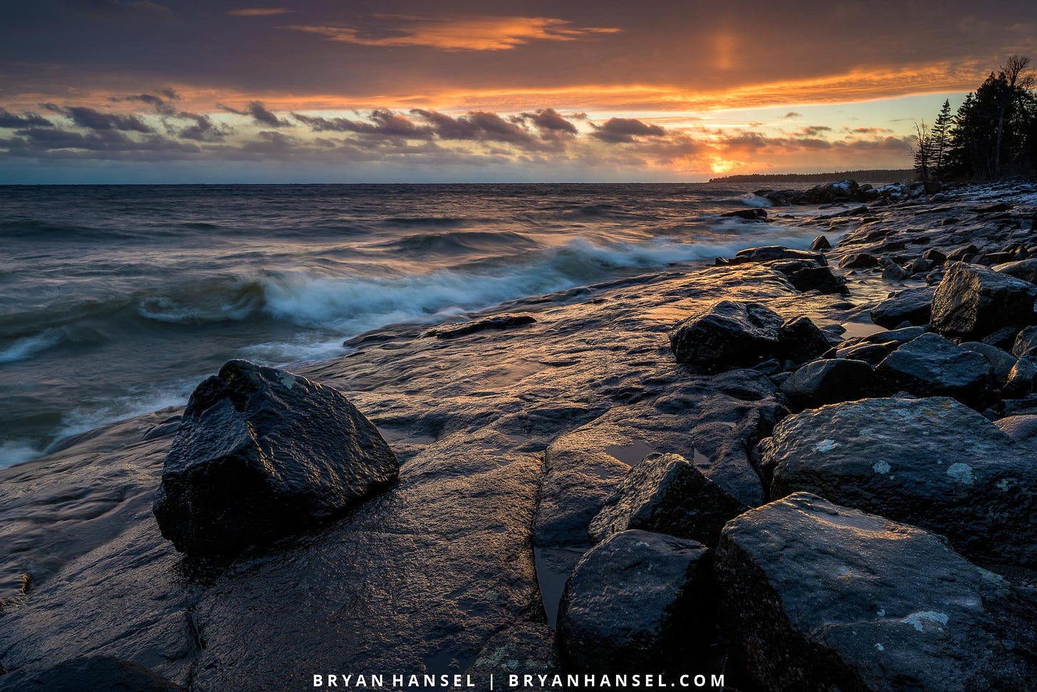 A photo of a basalt shoreline at sunset. The wet black surface reflects the red of the sunset. Lake Superior is rolling in waves on the left of the shot. An exercise ball sized rock is in the lower right.