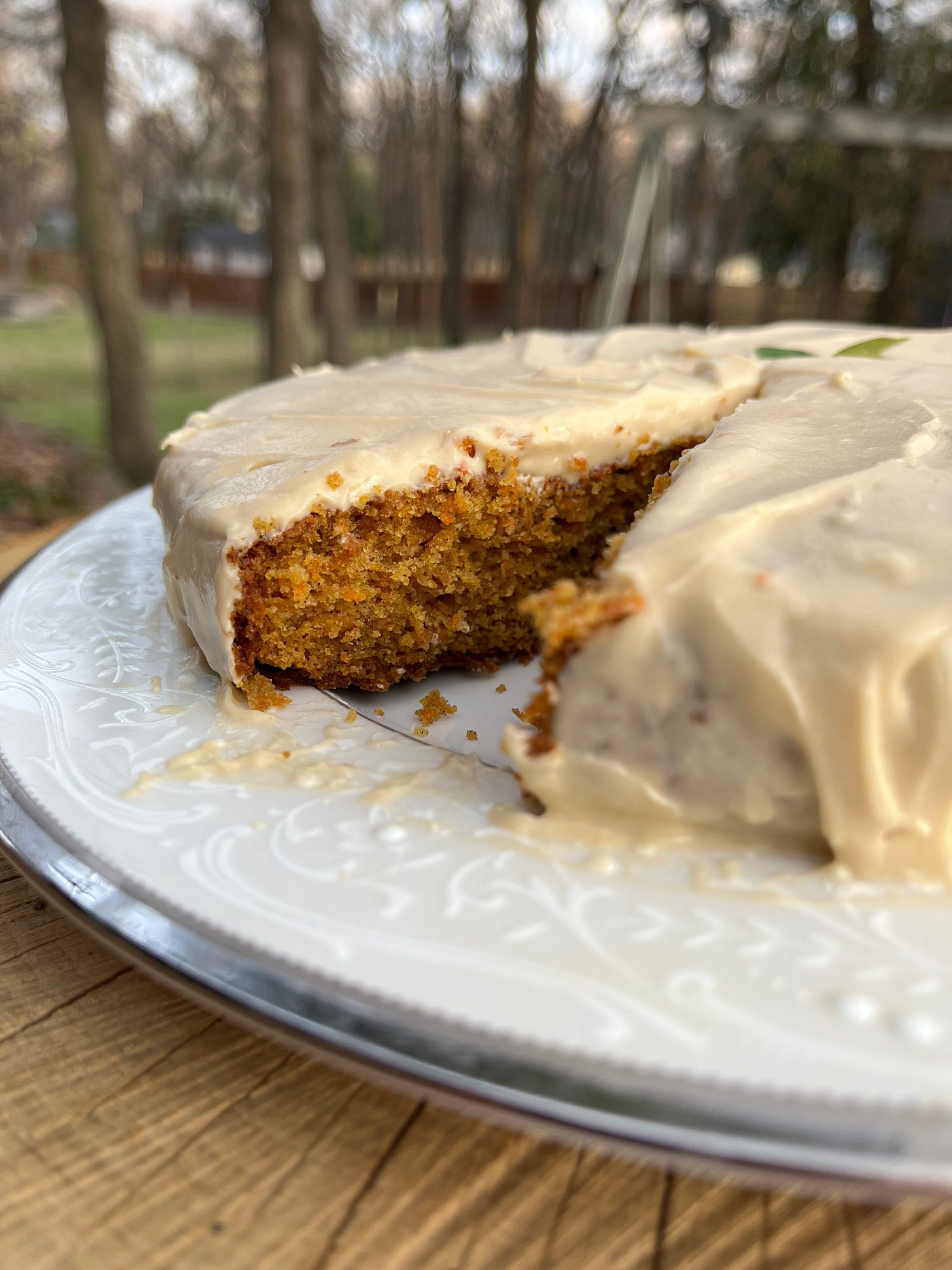 single layer carrot cake with cream cheese frosting, one slice removed, displayed on a white plate on a stump outside.