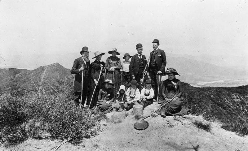 A group of formally dressed men and women pose on top of a hill, with more mountains and a river in the background dated 1885