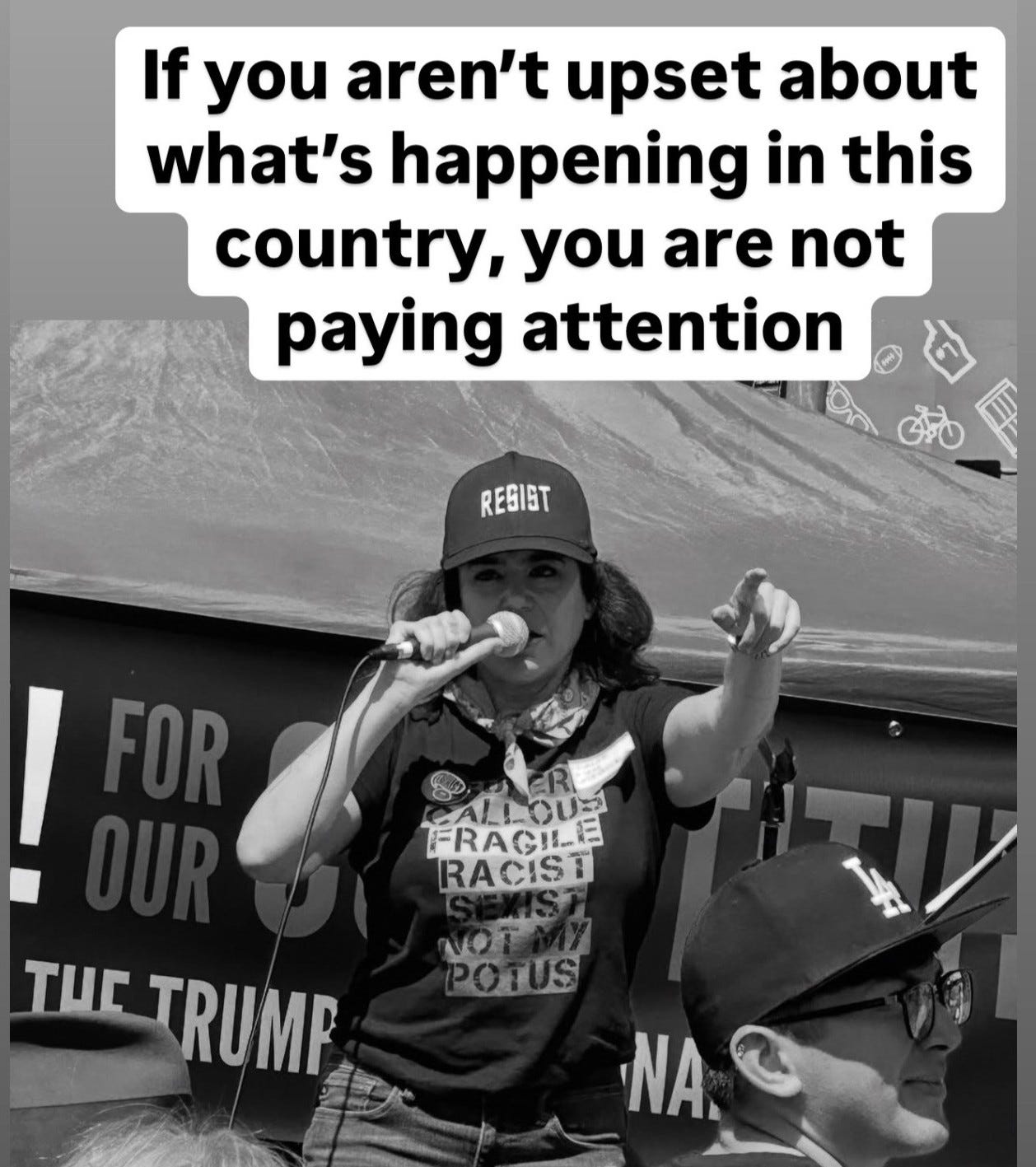 Text at the top reads "If you aren't upset about what's happening in our country, you're not paying attention." Below is black and white photo of a woman wearing a hat that says RESIST and a t-shirt that says Super Callous Fragile Racist Sexist Not My POTUS. She his holding a microphone and gesturing to the crowd in front of her. Behind her is a sign that says "March For Our Constitution." Text at the top reads "If you aren't upset about what's happening in our country, you're not paying attention." Below is black and white photo of a woman wearing a hat that says RESIST and a t-shirt that says Super Callous Fragile Racist Sexist Not My POTUS. She his holding a microphone and gesturing to the crowd in front of her. Behind her is a sign that says "March For Our Constitution."