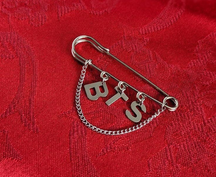 Silver safety pin with the letters “BTS” hanging from it by small loops and chain, resting on rich red patterned fabric. Symbol of what's often unseen but essential. Silver safety pin with the letters “BTS” hanging from it by small loops and chain, resting on rich red patterned fabric. Symbol of what's often unseen but essential.