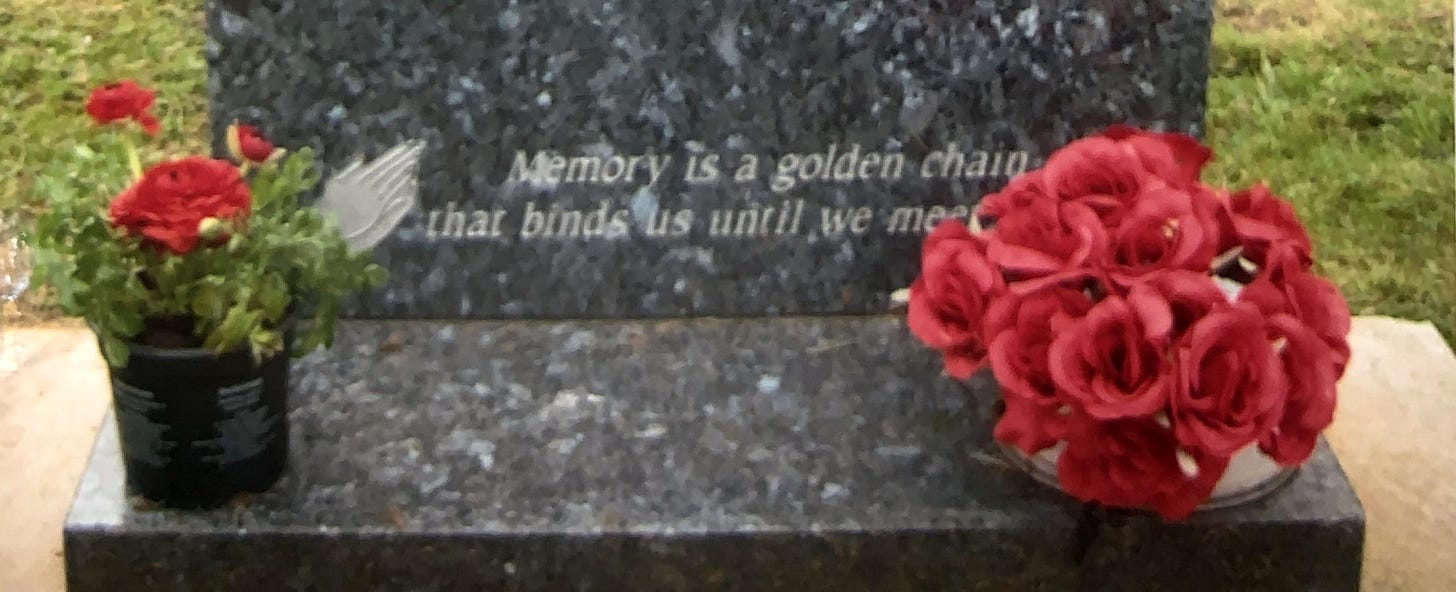 A gray grave with a pot of red flowers on the left and a bunch of fake red flowers on the right. Words on the grave are Memory is a golden chain that binds us until we meet again