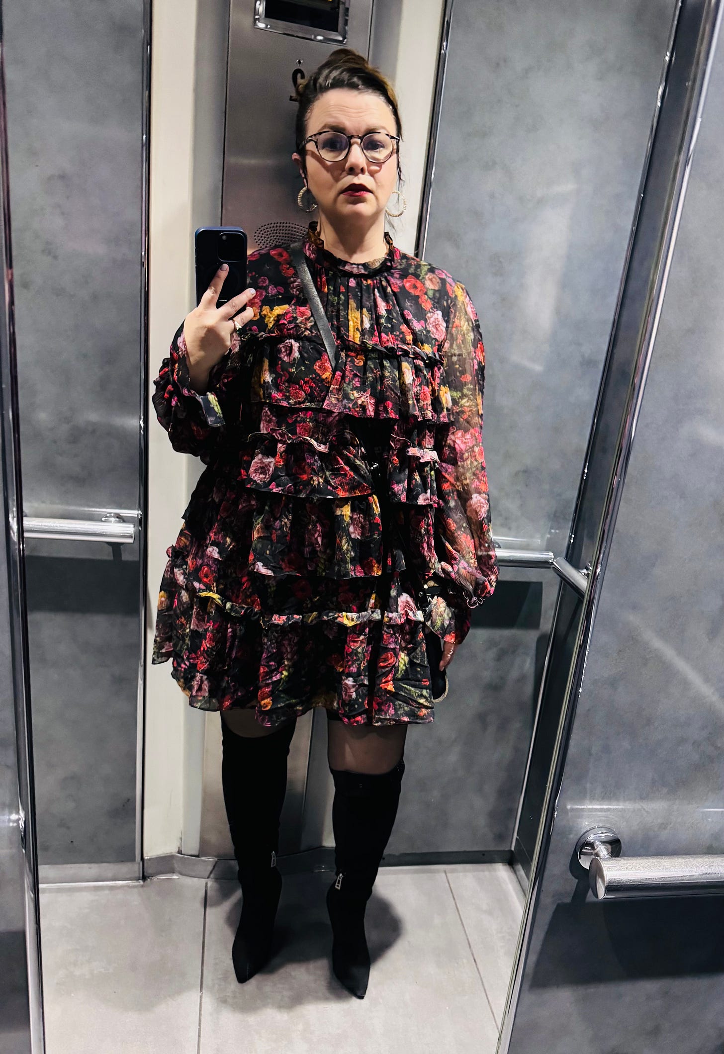 Amber takes a selfie in an elevator mirror wearing a long-sleeved, tiered dark flower print dress with thigh-high black boots. 