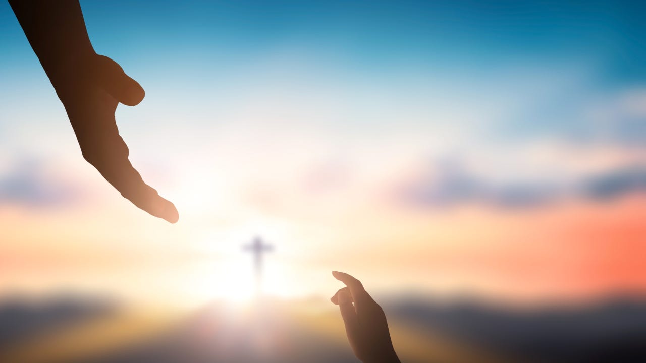 A person's hand reaching out to another person's hand with the cross in the background.