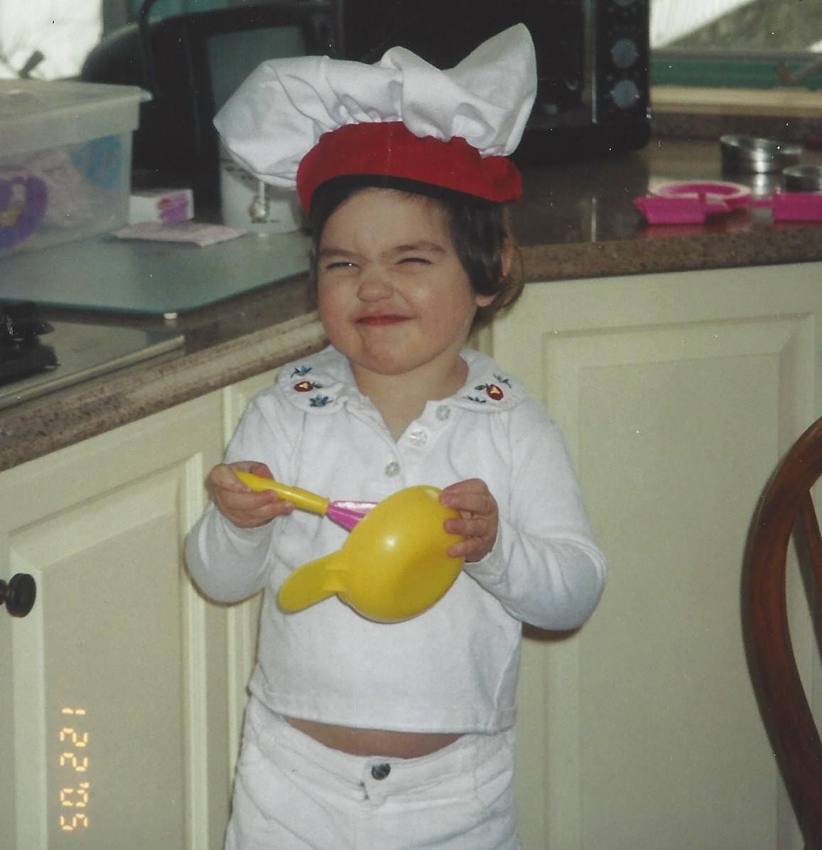 3-year-old girl wearing a mini chef's hat using a kid-sized utensil and bowl to whip something up in the kitchen