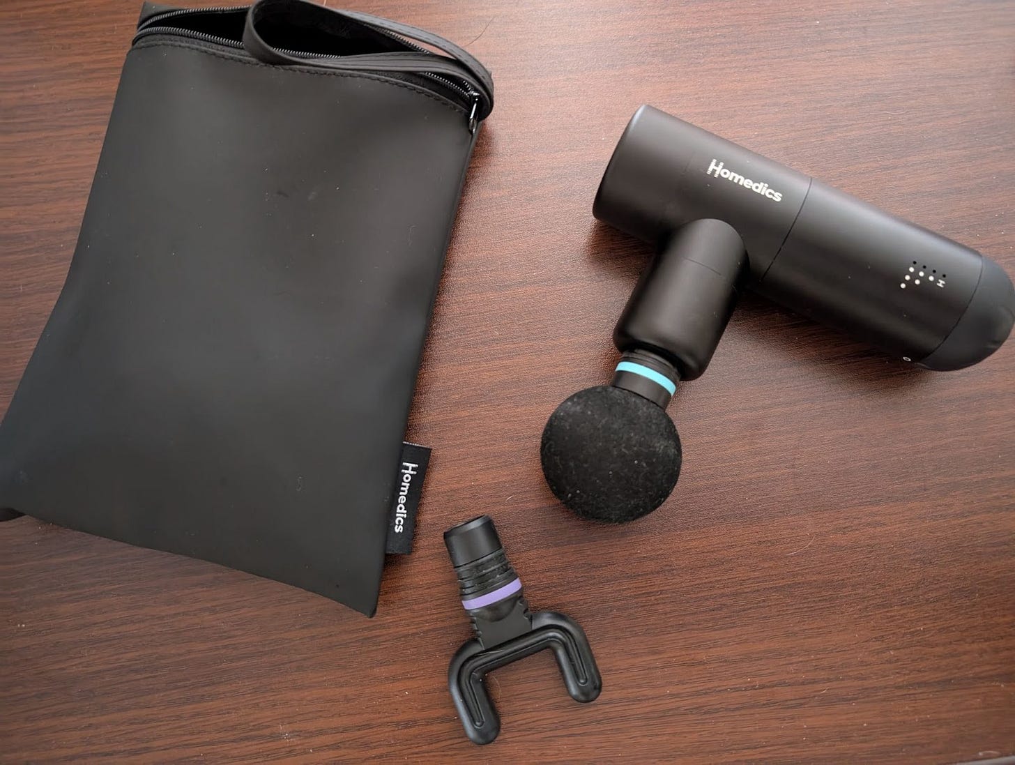 Author’s picture of Hometics portable massage device with a foam ball attached, lying on a dark wood surface beside the black case and two-pronged attachment.