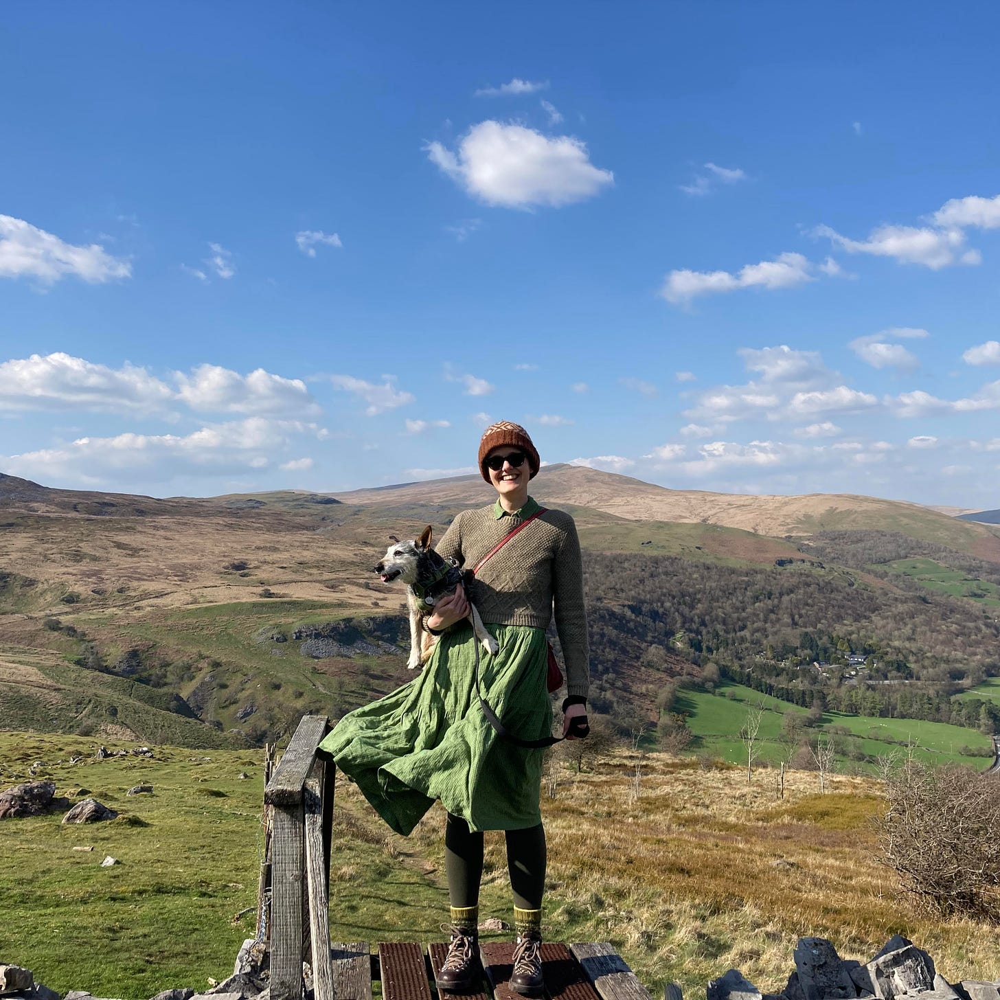  Katie the human stands on a stile halfway up a mountain, holding Jack the dog in their right arm, with a splendorous panoramic view of rolling hills and wooded slopes in the background. The sky is bright blue and speckled with fluffy white clouds. Katie is wearing a woolly hat, sunglasses, woolly jumper, green dress that is billowing in the wind, green leggings, woolly socks and walking boots. Jack is gazing off left towards the sheep.