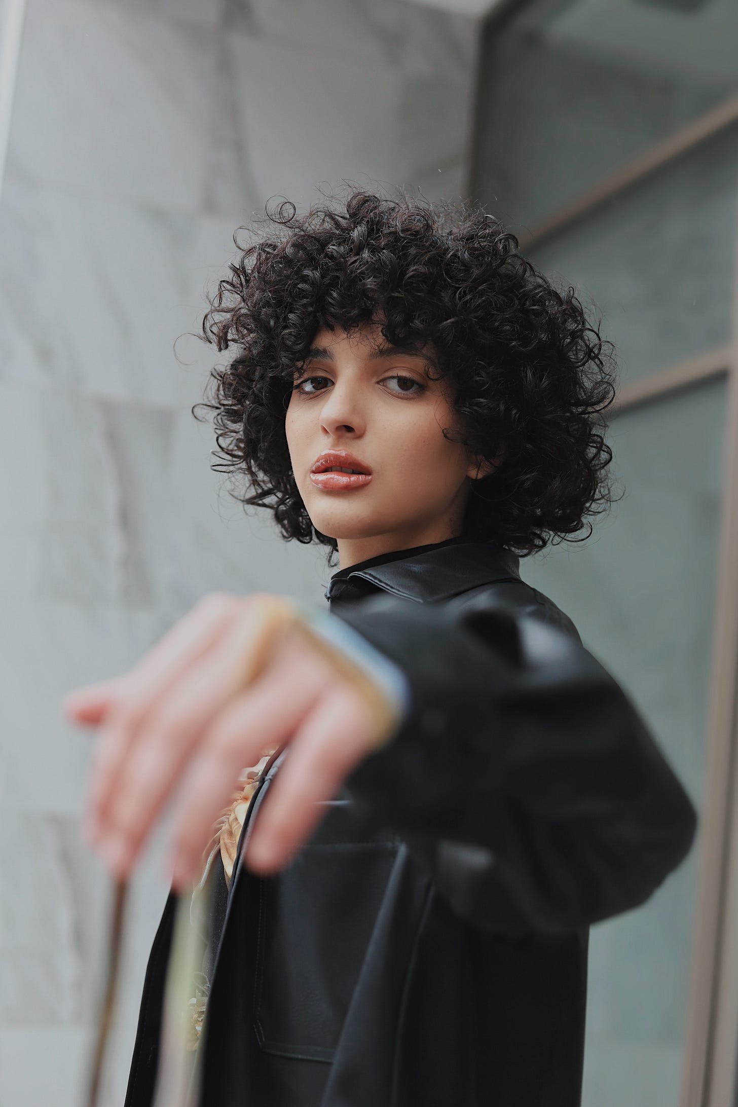 A young person with short, curly black hair looks at the camera with a straight face. They are wearing a black leather jacket.