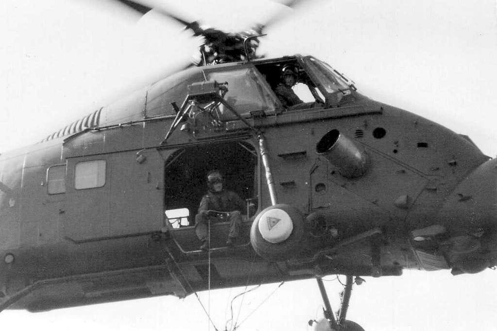 Black and white photo of the side of a Wessex helicopter. The pilot and winchman can be seen. It is unclear how they got so many people into a relatively small cargo bay.
