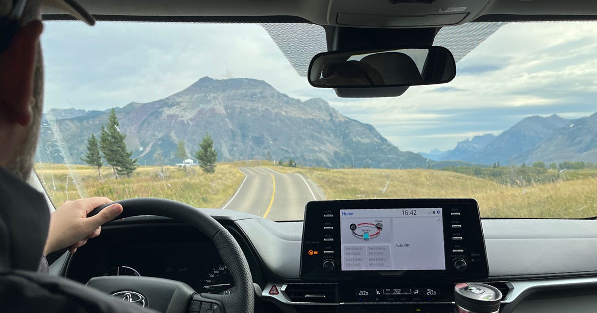 View out the front window of our accessible van. Over the dashboard and the steering wheel, the road stretches across the golden-brown prairies spotted with green trees. In the distance, blue-grey mountains rise up and stretch to the horizon.