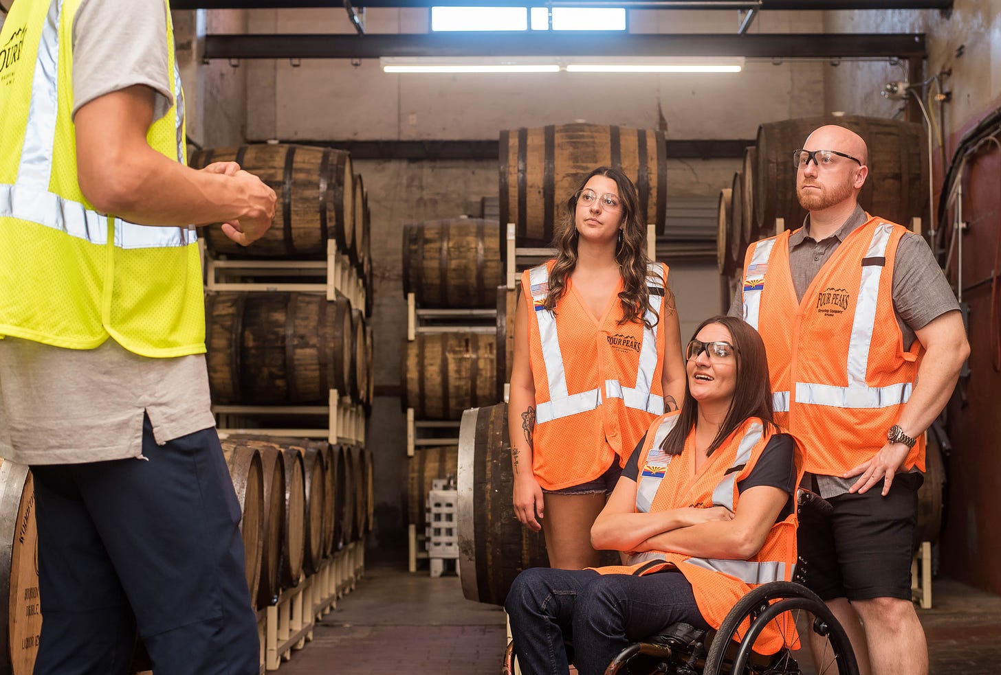 Three people in orange high-vis vests, one woman is in a wheelchair, looking at someone in a yellow vest. In the background are barrels.