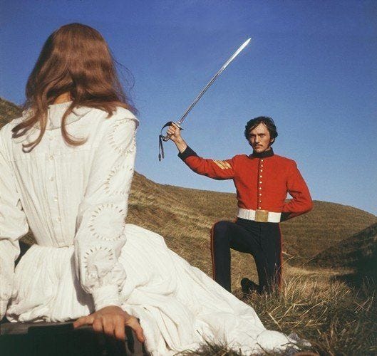 film still of a woman in a white dress looking a bit taken aback in front of a man in military garb brandishing a sword