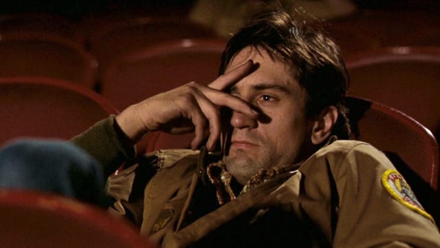 A screenshot from Taxi Driver showing Travis Bickle slouched in a movie theater seat with this hand partially covering his face.