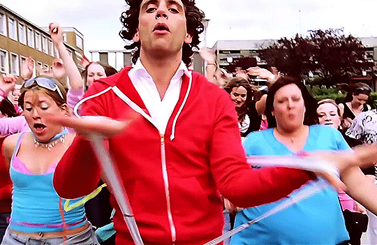Gif of Mika with curly brown hair and a red hoodie dancing down the street, surrounded by women