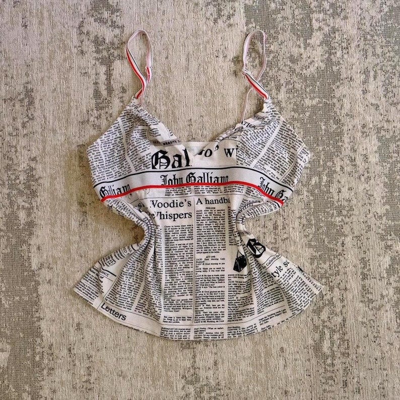 May include: A white sleeveless top with a newspaper print design. The top has a red and white stripe across the chest with the text 'John Galliano' printed on it. The newspaper print includes the text 'Gal '70's', 'A handbag', 'Hoodie's Whispers', 'Letters', and 'Style & Substance'.