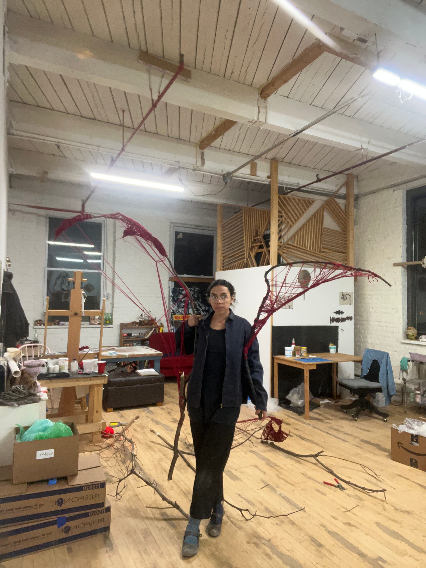 i am standing in the middle of a friends art studio holding two of the scepters i made. found tree branches wrapped in red yarn the way i am holding the mimics wings. 