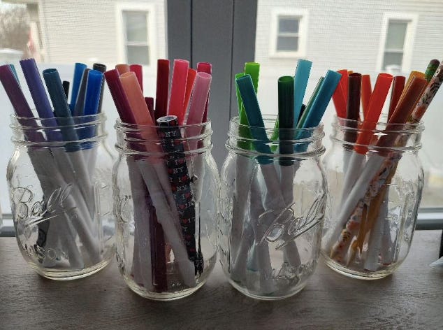 Four mason jars, each filled with markers, line a windowsill.