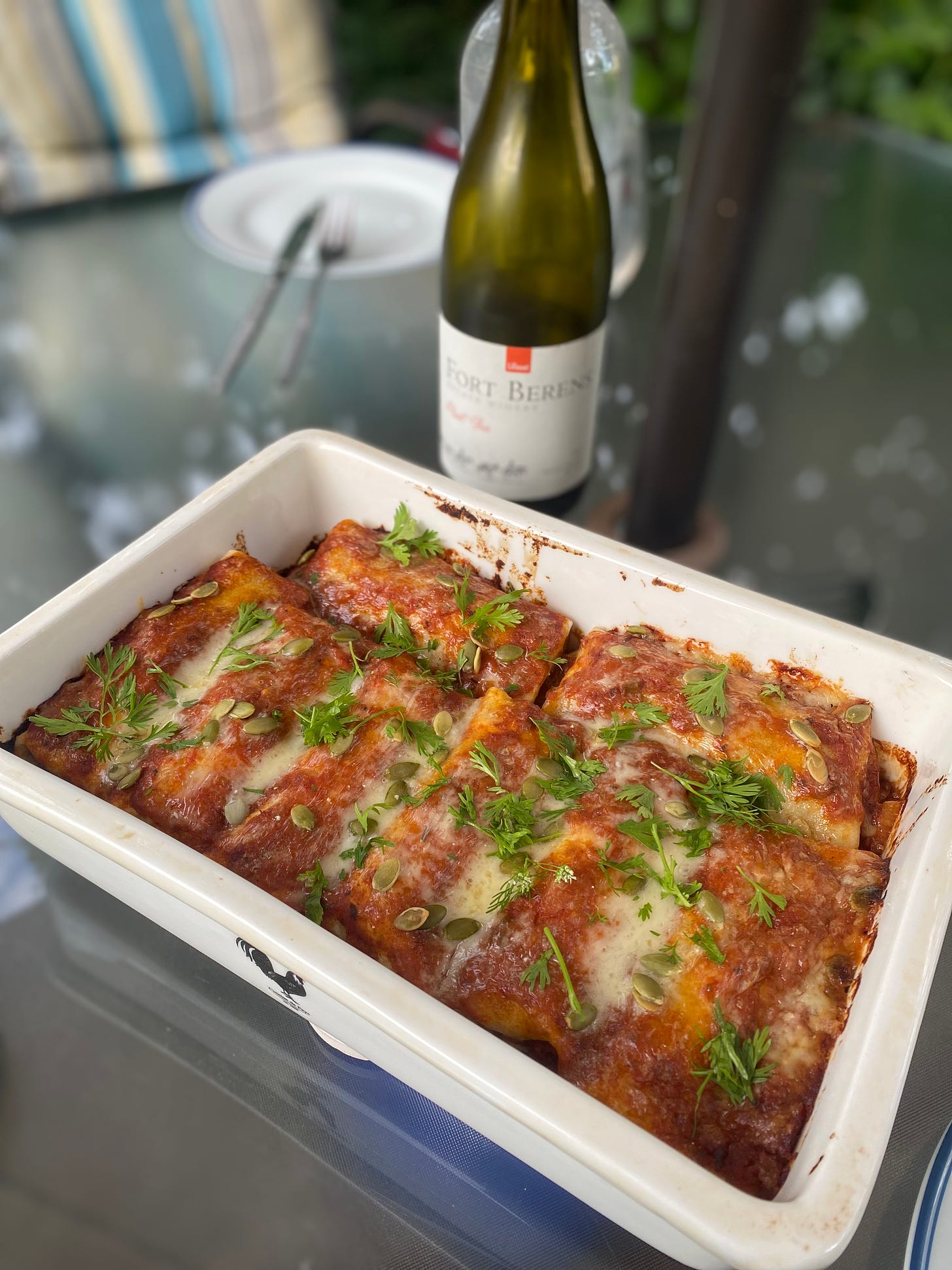 A white rectangular dish of enchiladas in red sauce, melted white cheddar over top. Sprinkled across it are pepitas and cilantro. In the background is a bottle of Fort Berens pinot gris.