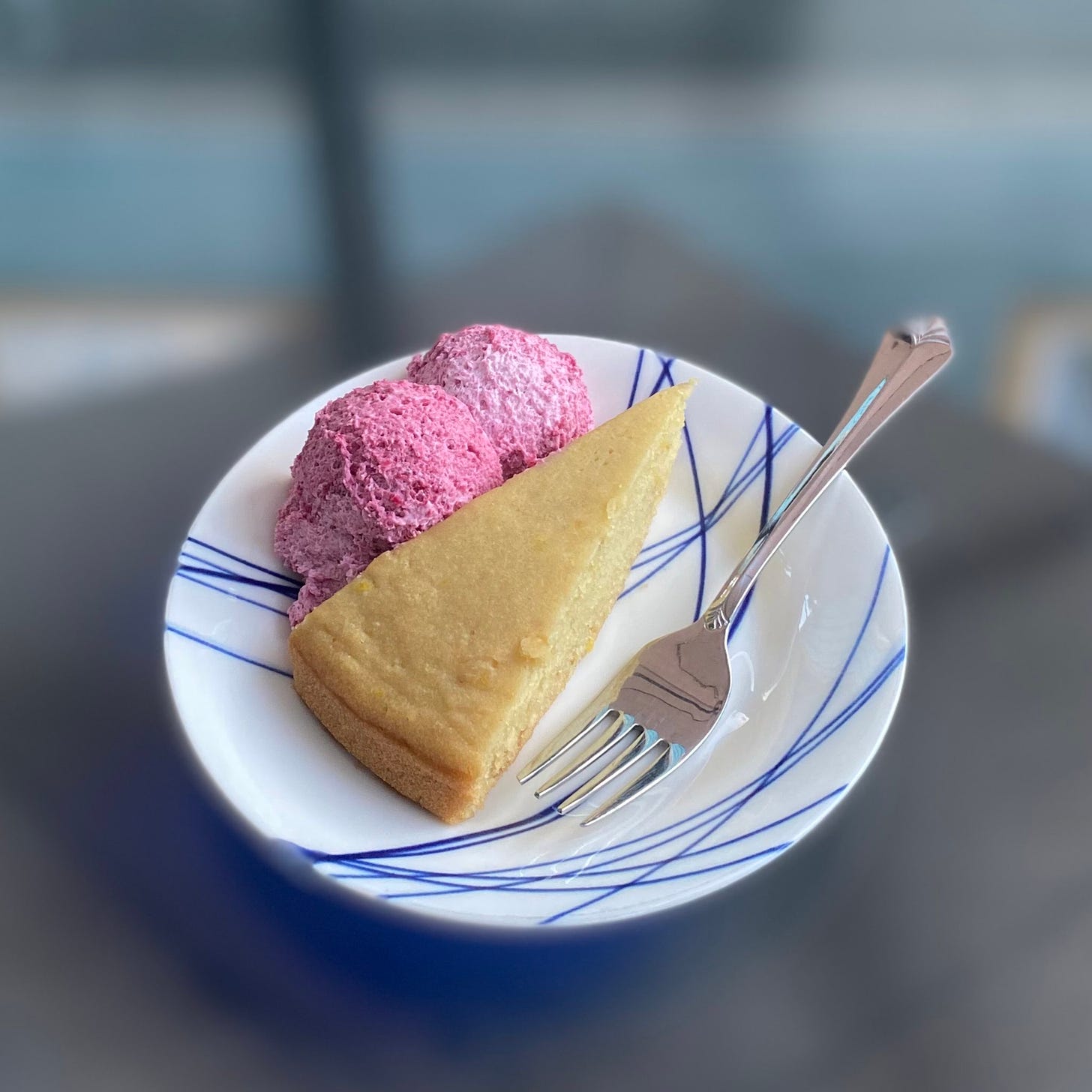 A small blue and white plate with a slice of olive oil cake, a fork in front of it. On the other side of the cake are two small scoops of deep pink strawberry Bavarian cream.