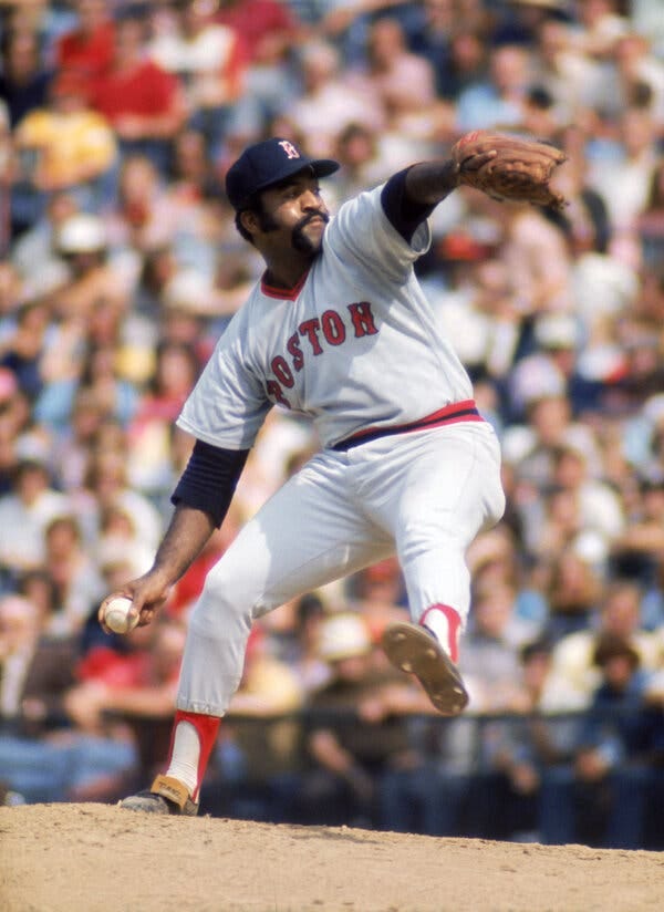 Luis Tiant, an intense-looking man with a long mustache, standing on the pitching mound and prepares to through. His left leg is extended, his left arm is lifted high, and his right arm hands at his side.