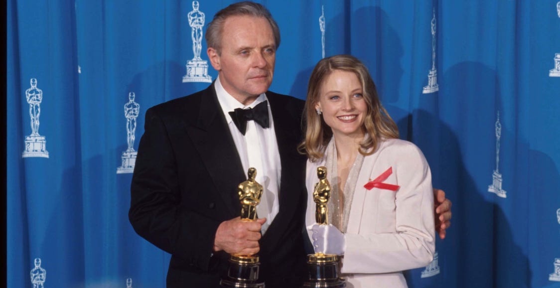 Fotografia de Anthony Hopkins ao lado da atriz Jodie Foster - cada um com uma estatueta do Oscar nas mãos. Ele é branco, alto, tem cabelo grisalho, olhos azuis e usa smoking; ela, branca, loira com cabelo na altura do ombro, com terninho rosa claro. 
