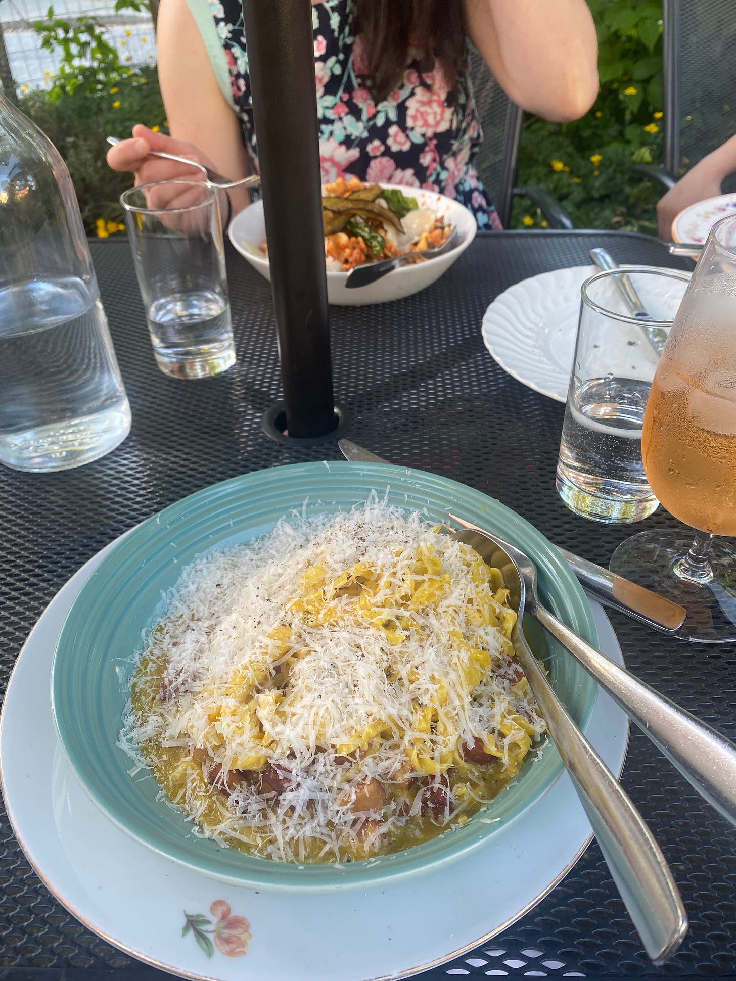 A powder blue bowl of carbonara pasta with lots of grated parmesan on top. It rests on an old-fashioned china plate, white with a tulip design. An aperol spritz is just visible in the frame next to it, and across the table, Natalie's fork hovers over her bowl of eggplant pasta.