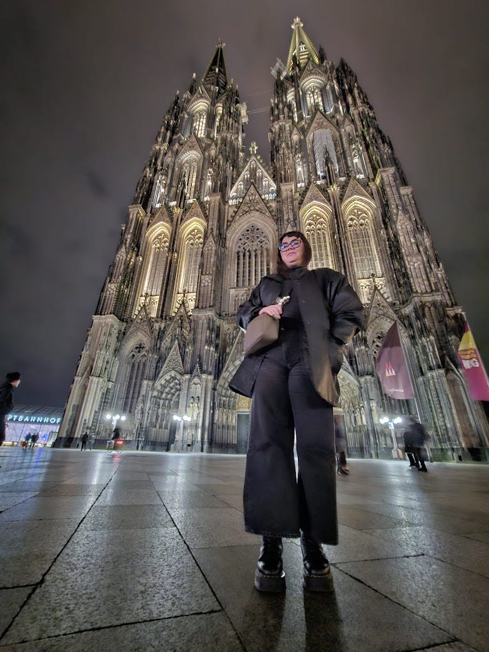 Me, wearing black, stood in front of Cologne Cathedral at night. The windows are lit in gold, and the stone looks grey and black. The photo is taken from the ground.