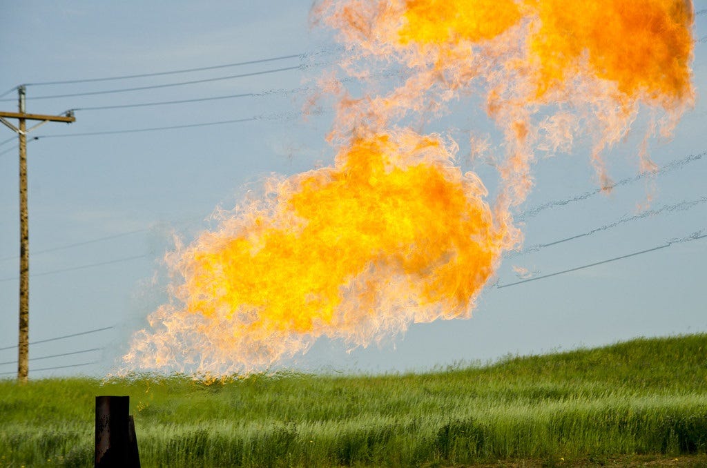 Photo of gas being burned from an oil well. Bright orange-yellow flames stand out against a pale blue sky and green farm fields.