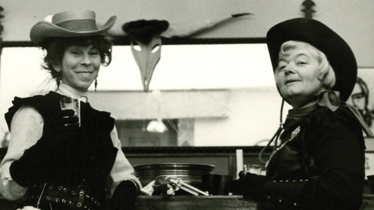 Two women stand leaning against a bar in Western garb, holding drinks in their hands. They are bother smiling. A steer skull with horns is on the wall behind them and there is an empty and untouched revolver between them for prop purposes. The photo is black and white and painfully adorable.