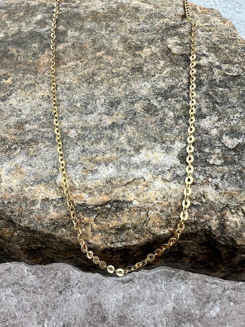 May include: A gold chain necklace with small, round links. The chain is laying on a textured, gray rock surface.