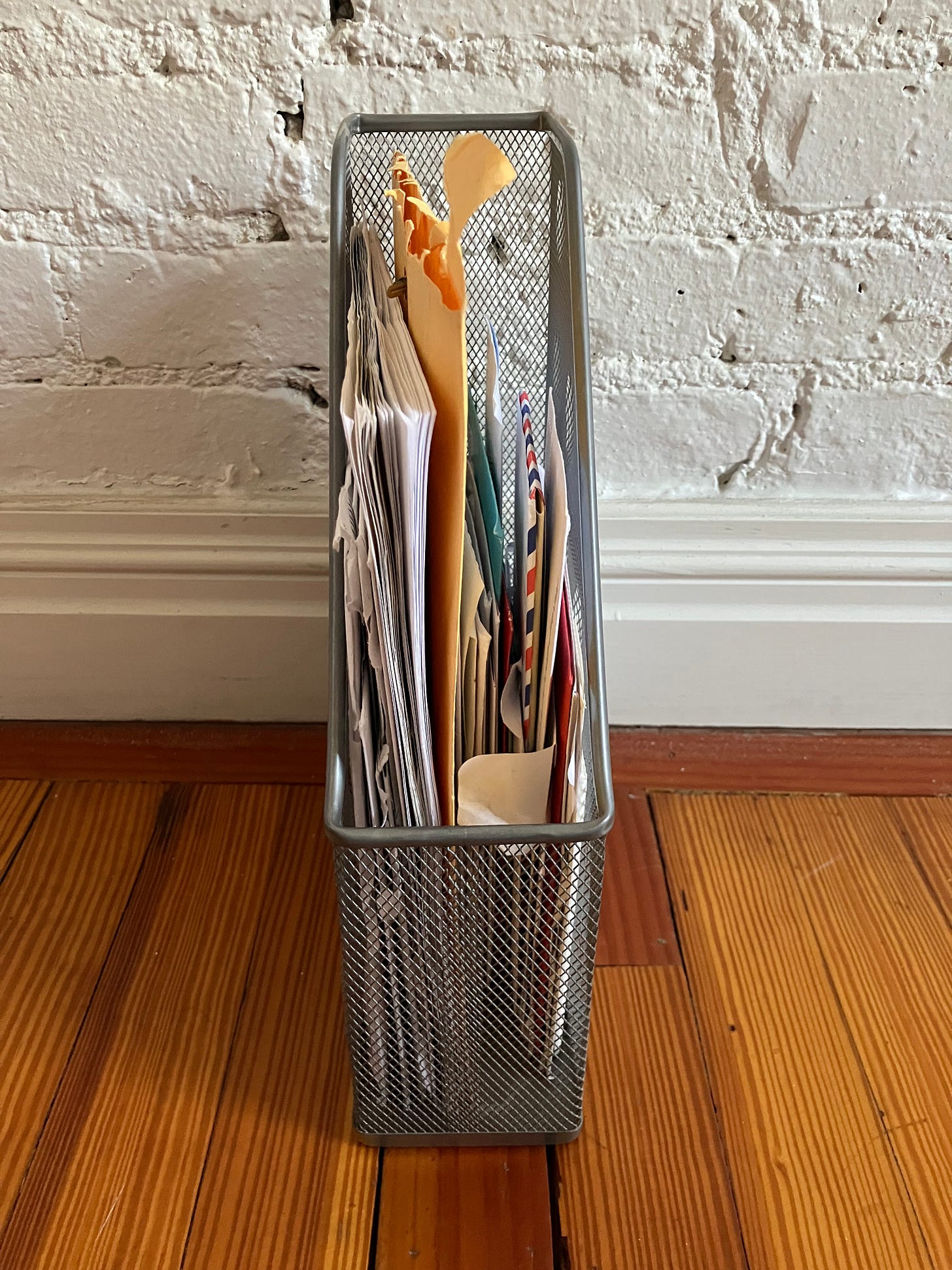 a wire basket filled with assorted letters and envelopes on a bare wood floor against a white brick wall