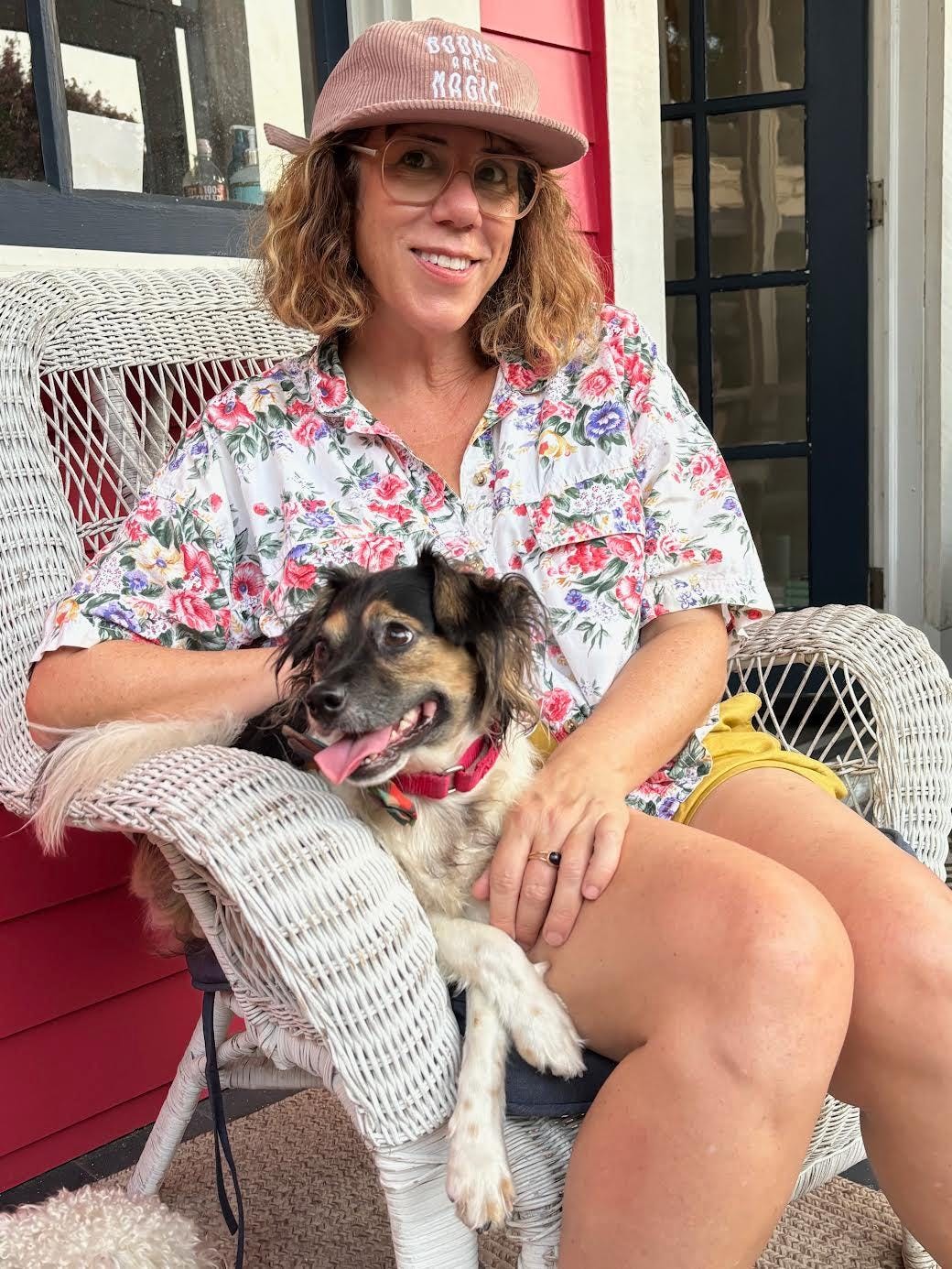 woman with salmon colored baseball cap in white wicker chair with small mixed breed dog which has a red collar and tongue hanging out woman is writer jami attenberg