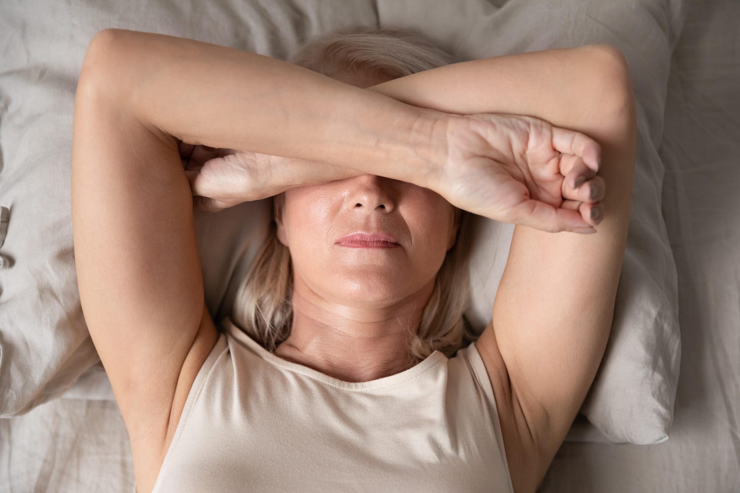 Mature woman laying in bed with arms over face suffering from insomnia 