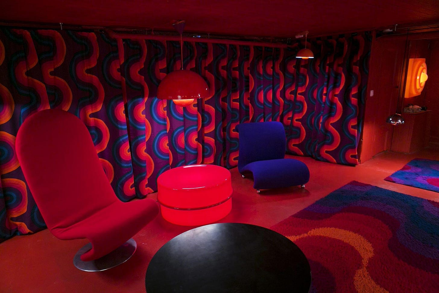 Photo of a 1970’s interior with modern armless armchairs, thick curtains and carpets characterised by rich and deep reds, blues, and purples.