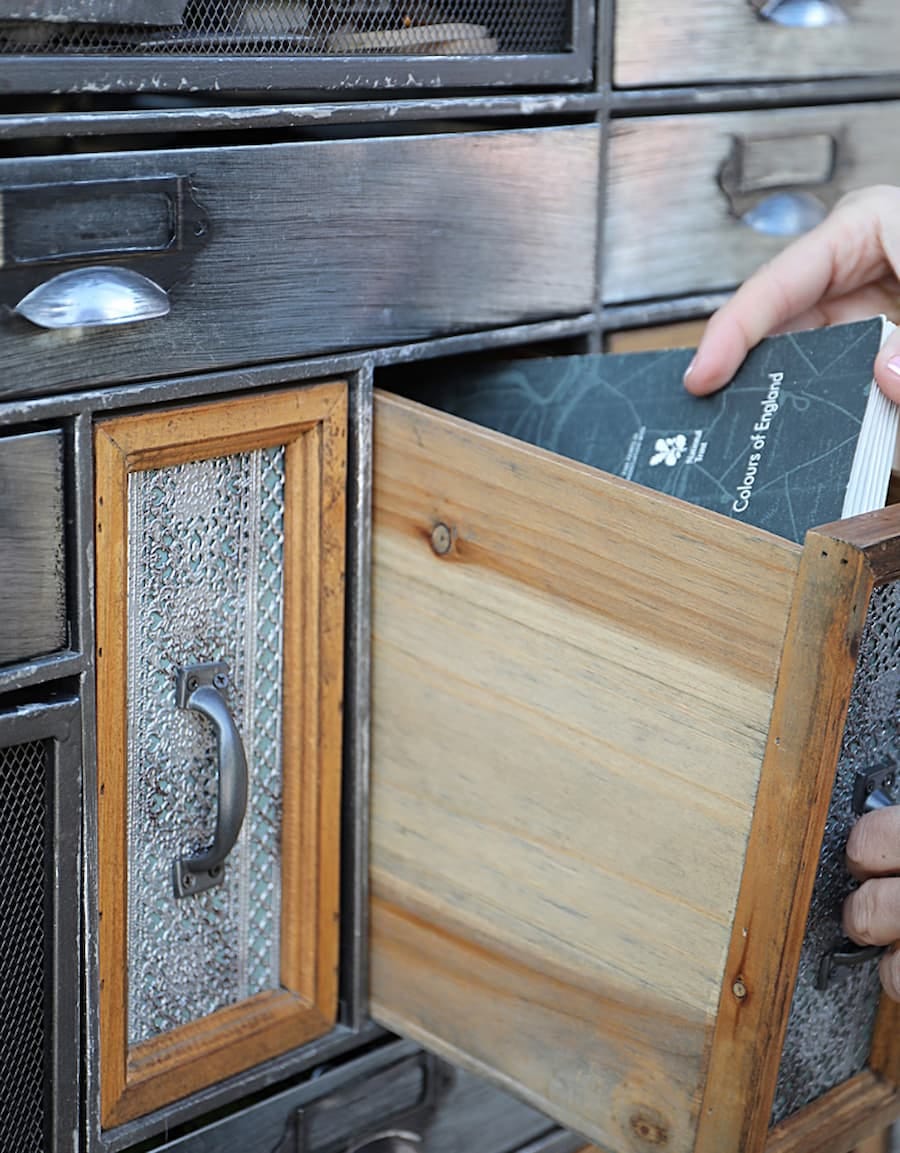 A hand pulling out a drawer from a wooden cabinet. The cabinet has multiple drawers with different designs and textures. The drawer being pulled out is made of wood and has a colour chart with a dark grey cover being pulled out.