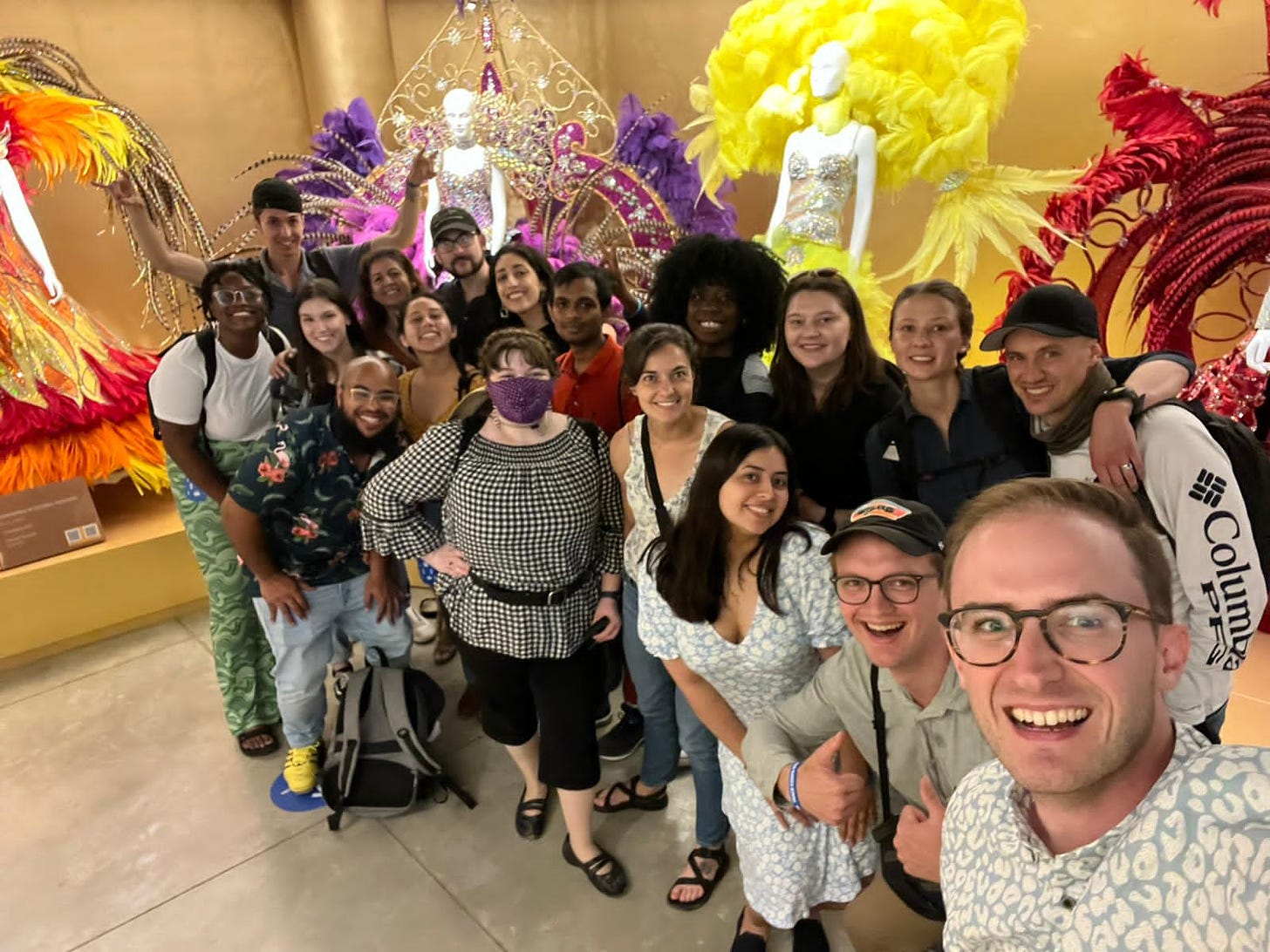 a group photo of 18 people standing in front of mannequins dressed in carnaval costumes