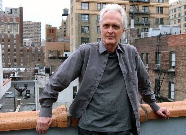 Scott Johnson, an older man with white hair wearing a gray shirt and sweater, leans back against the railing on the roof of a New York City apartment while looking at the camera with a half-smile on his face.