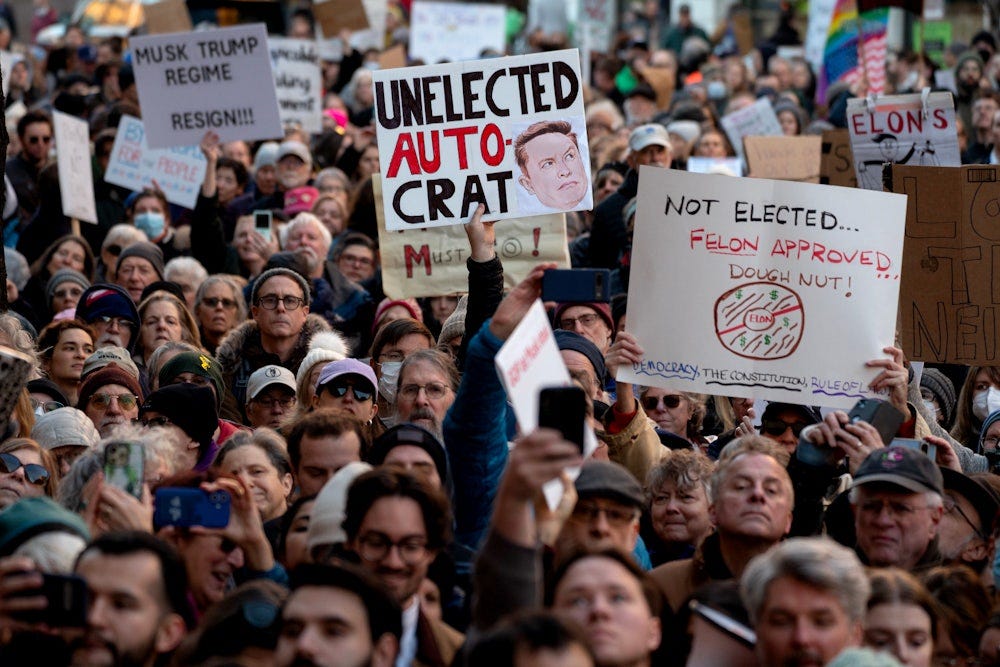 Demonstrators face the camera holding signs reading, among other messages, "Unelected Autocrat" and "Musk Trump Regime Resign!"