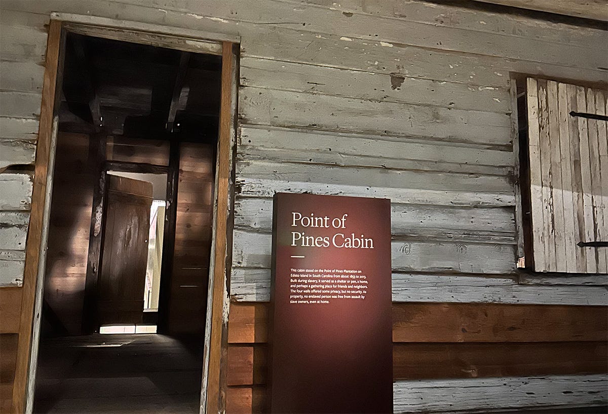 A blue/gray wooden frame house with open doors and a red brick foundation. A placard next to it labels it as the Point of Pines Cabin.