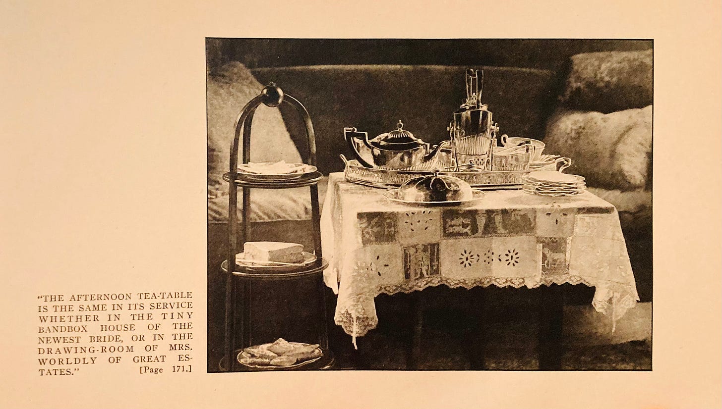 Image from 1922 of an Everday Afternoon Tea Table. tiered tray with cakes, table with elaborate table cloth, silver tea set with stack of plates. There is a velvet couch in the background. Photo is black and white with a sepia tone quality to it. 