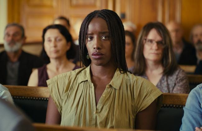 A woman in braids and a summery olive green shirt is seated amongst other observers in a courtroom. She has an intent look in her eyes.