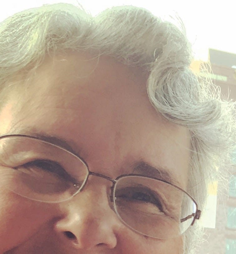 A gray-haired, bespectacled woman with a big curl on the left side of her forehead.