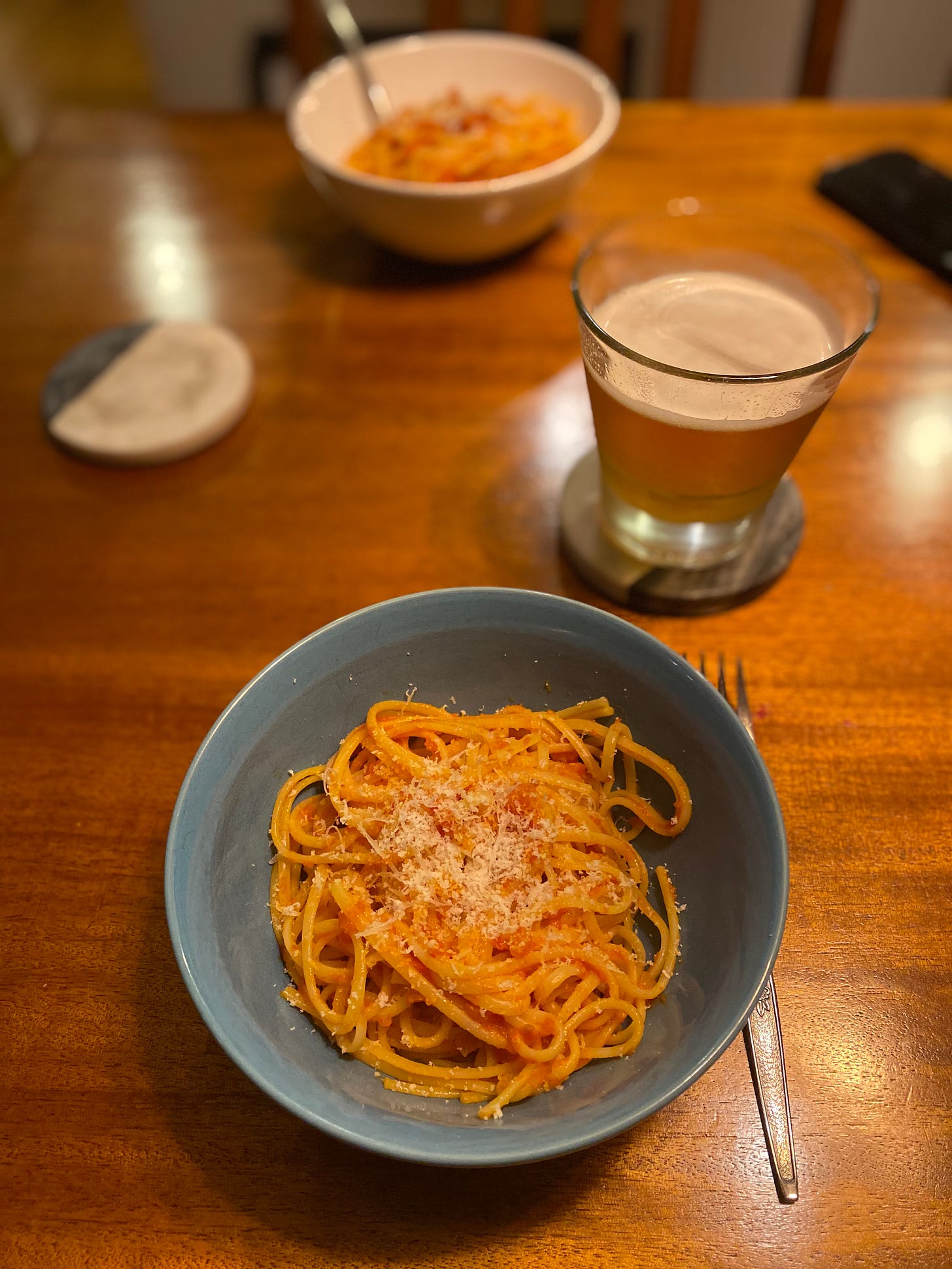 A blue bowl of pasta in tomato sauce, with parmesan cheese on top. On a coaster behind it is a glass of pale ale, and in the background is a white bowl of the same pasta.