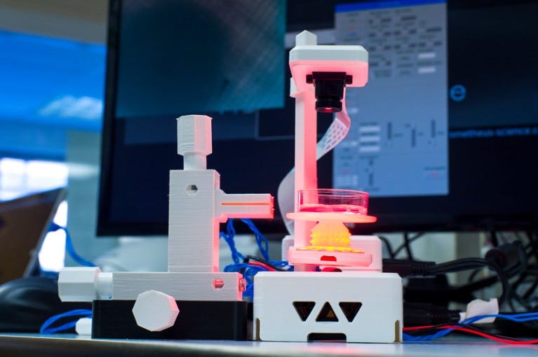 A 3D printed fluorescence microscope with lens looking down onto a petri dish is set up on a lab bench with a monitor behind