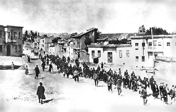 A long column of people leaving a town. Alongside the column, men with rifles are marching with their rifles ready.