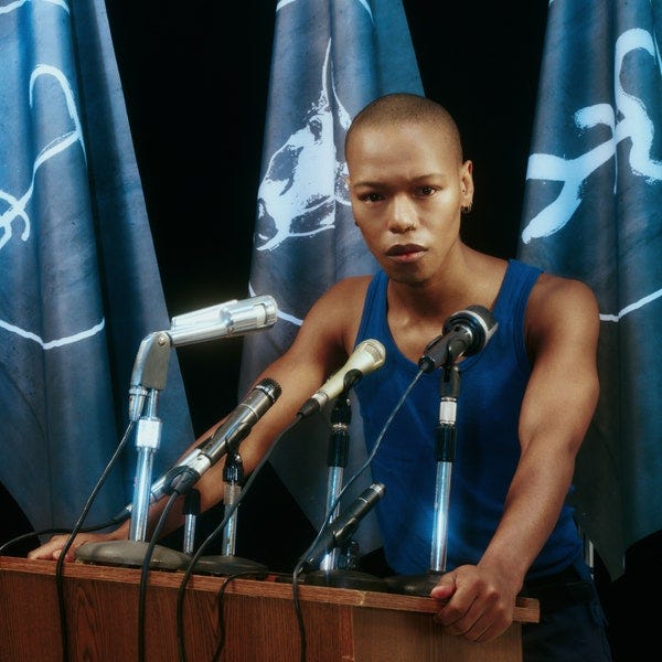 Nakhane, wearing a tank top, stands behind a podium with several microphones on it.