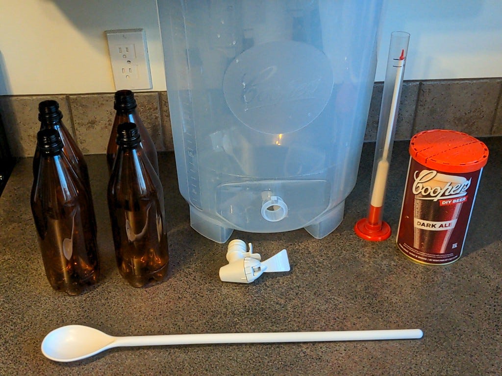 components from a beer brewing kit lay spread out on a kitechen counter. they include a large plastic tub, some empty clean plastic bottles, a stirring spoon, a hydrometer, and a can of dark malt extract.