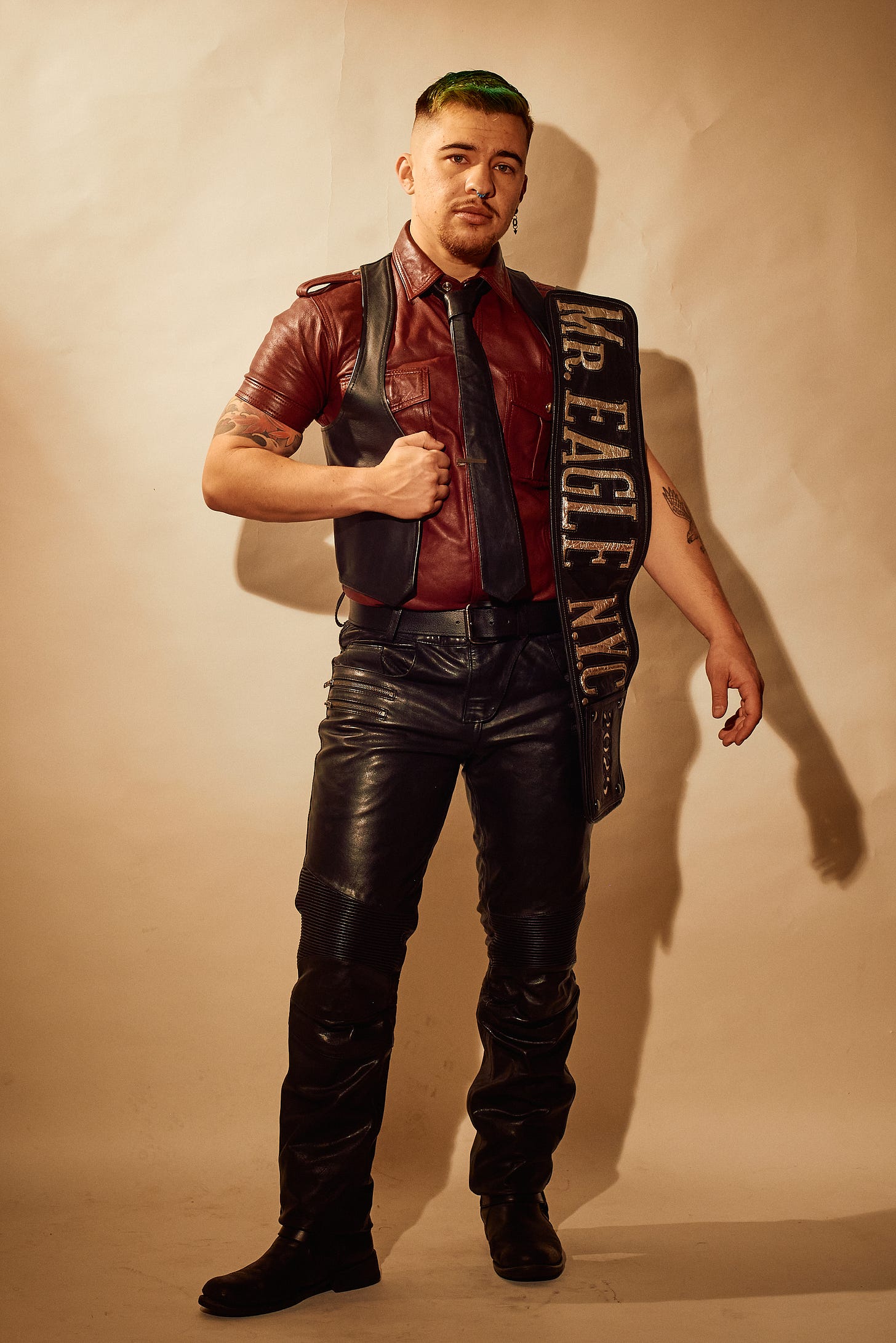 A person stands confidently in leather gear, wearing a red leather shirt, black tie, and black leather pants with boots. They have green hair, tattoos, and are holding a “Mr. Eagle NYC” sash over their shoulder, exuding a bold and assertive presence.