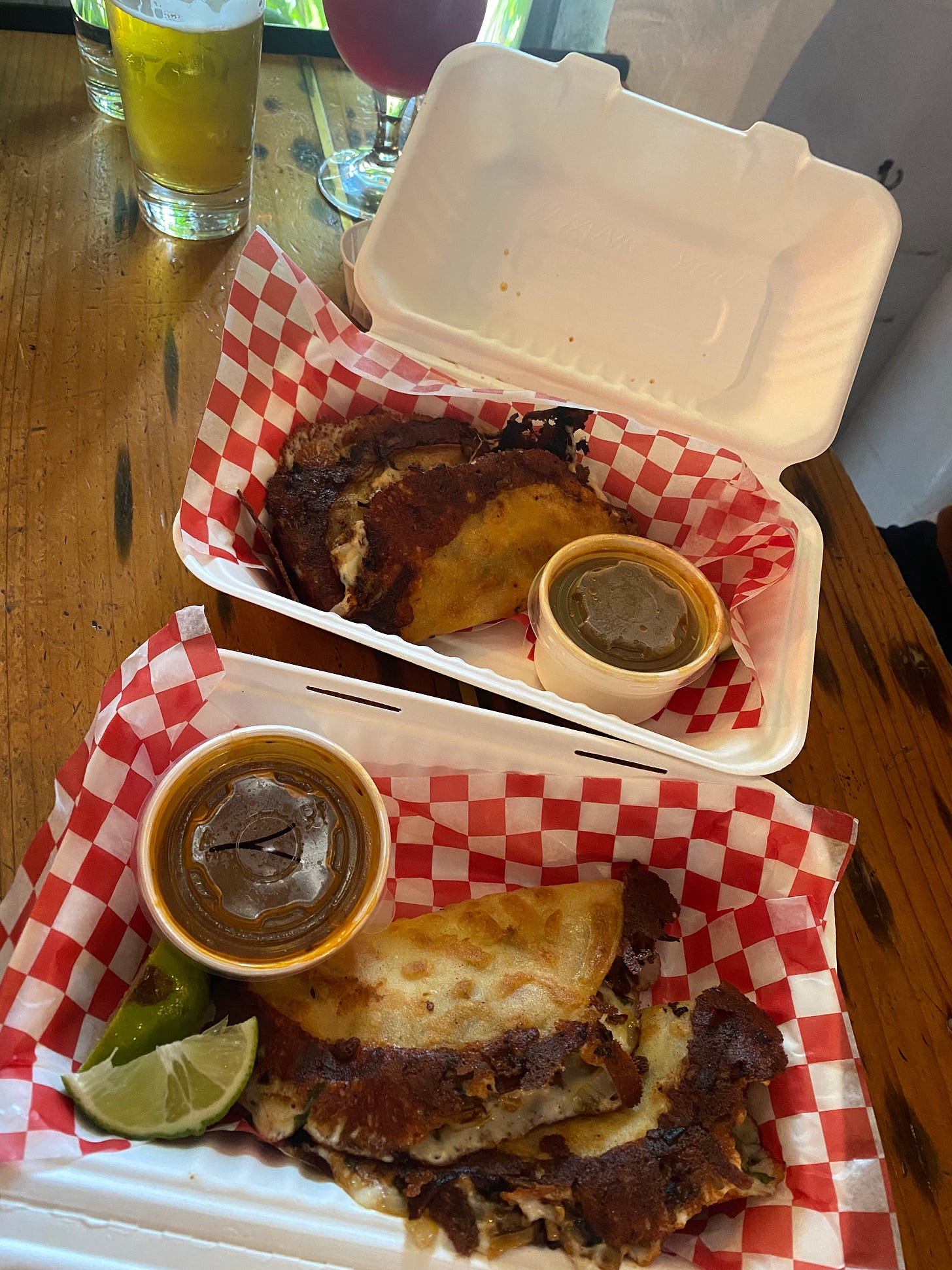 White paper takeout boxes lined with red and white checkered paper, with two birria tacos each, slices of lime and small containers of sauce next to them. In the background are two glasses of beer, one pale and one reddish-purple.