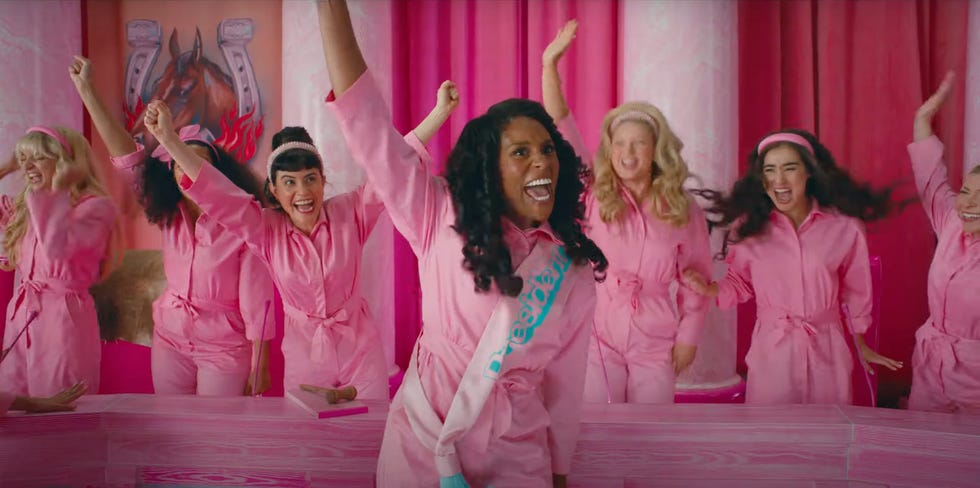 A Black woman wearing a pink jumpsuit and a lighter pink sash with blue lettering that reads President, raises her arm up as she cheers and smiles. She is flanked by six other women also in pink jumpsuits and cheering, while in a pink-clad room.